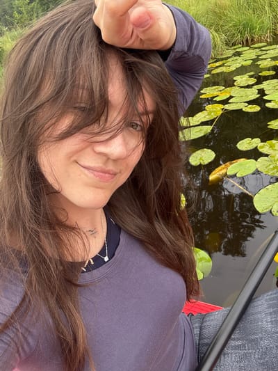 A person with long, brown, layered hair is taking a selfie in a body of water with lily pads. The person is wearing a purple long-sleeved shirt and a necklace. Their right hand is raised and resting on their head. A paddle is visible to the right of the person. The background consists of tall green grass and the dark water with lily pads. The location is the city of Mesliki, Russia.