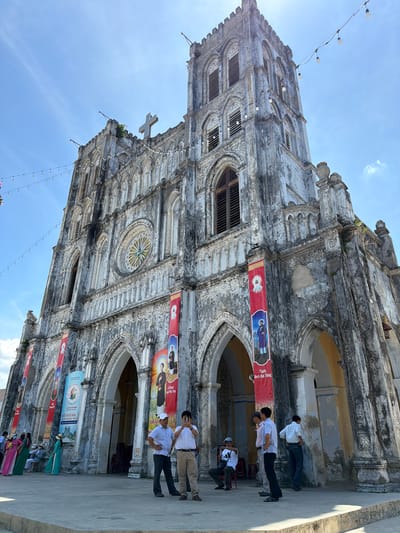 The image captures a low-angle view of an old, weathered church in Hội Tín, Vietnam, bathed in bright sunlight under a clear blue sky. The church's facade is aged, with peeling paint and visible signs of wear, contributing to a historic and somewhat eerie atmosphere. A prominent cross sits atop the central tower, and decorative string lights are strung across the sky, adding a festive touch.

Several red banners with images of religious figures hang vertically on the church's exterior. In the foreground, a group of people are gathered on the paved area in front of the church. Some are posing for a photo, while others are engaged in conversation. The people are dressed in casual clothing, and some are wearing hats. The overall scene suggests a visit to a significant cultural or religious landmark.
