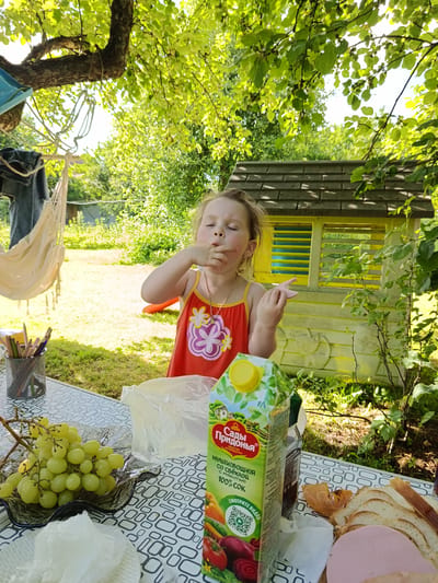 In Shishkin Les, Russia, a young girl with blonde hair tied back is shown with her eyes closed, eating with her right hand while sitting at a table outdoors. She is wearing an orange sleeveless dress with a floral design. The table is covered with a patterned cloth and holds a bunch of green grapes in a dark glass dish, a plastic bag, a juice carton with Russian labeling that says "Сады Придонья" and "Мультиовощной со свеклой 100% сок," a bottle of what appears to be oil, and sliced bread with a slice of ham. In the background, a yellow playhouse with a dark roof is visible, surrounded by greenery. To the left of the girl, a hammock is hanging, and a mesh container with colored pencils is on the table. The scene is set in a garden with dappled sunlight filtering through the leaves of a large tree.