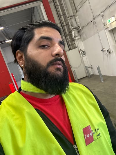 The image shows a close-up, selfie-style shot of a man in a warehouse or industrial setting. He has dark hair, a full beard, and is wearing a red t-shirt and a bright yellow safety vest with a "logLine express" logo. The background is industrial, with concrete floors, metal pipes, electrical conduits, and an emergency exit sign. The man appears to be looking directly at the camera with a neutral expression. The lighting is typical of an indoor industrial environment. The presence of the safety vest and the warehouse setting suggest he is likely working there. Given the "logLine express" logo, this could be a logistics or distribution center.