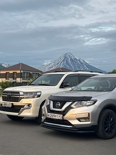 In Razdol'nyy, Russia, two SUVs are parked on a paved road with a residential building and a snow-capped volcano in the background. The white SUV on the left, a Toyota Land Cruiser, has its front license plate visible, reading "C800CK 41". The silver Nissan SUV on the right has its front license plate reading "B269PT 41". The sky is overcast with scattered clouds. The residential building features a brown roof and a facade with dark and light stripes. The volcano, a prominent natural feature, dominates the background with its snow-covered slopes.