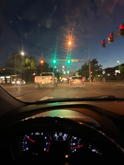The image captures a night view from the driver's seat of a car at an intersection in Pasco, Washington, USA. The dashboard, including the tachometer and speedometer, is visible in the foreground. The windshield shows a street scene with traffic lights, vehicles, and buildings. Traffic lights are illuminated green and red. A white pickup truck is in the immediate vicinity, and other vehicles are visible further down the road. The sky is dark with hues of blue and orange from streetlights and the setting sun. Trees and streetlights line the road. The overall impression is of a typical urban night driving scenario.