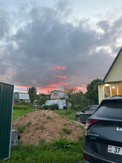 The image captures a rural scene in the city of Klyobushnevo, Russia, at dusk. A vibrant sunset with bright pink and orange hues is visible in the stormy grey clouds. In the foreground, a large pile of sand and green grass dominate the ground. To the right, the rear of a dark gray Kia Sportage is parked, with its license plate displaying "AM 37". Behind the Kia, a dark sedan is visible. A yellow house with a green roof is situated on the right side of the frame, with lights on inside its windows. In the background, several small houses with metal roofs, trees, and a greenhouse are scattered across the landscape, along with power lines stretching across the sky. A green metal fence is partially visible on the left side of the image.