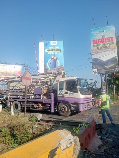 This image captures a street scene in Banjarkemantren, Indonesia, under a clear blue sky, suggesting a sunny day. The main subjects are a large concrete mixer truck, a person in a high-visibility vest, and two billboards in the background.

The concrete mixer truck, painted purple and white, is parked on a road, possibly at a crossing, indicated by the presence of a red and white striped barrier pole. The truck itself has a number "39" visible on its side. The person in the foreground, wearing a bright yellow vest with reflective stripes and blue jeans, appears to be directing traffic or observing the scene. They are holding a black bag and a small bucket.

In the background, two large billboards are prominently displayed. The left billboard, in blue, features the text "CIP KAWASAN PERGUDANGAN NOMOR SATU," which translates to "CIP Warehousing Area Number One." The right billboard, with a sky and cloud motif, advertises "THE BIGGEST INDUSTRIAL PARK IN SIDOARJO" and mentions "EAST JAVA INDONESIA." It also includes a warning section and a "21+" age restriction.

Other visible objects include road barriers, vegetation, streetlights, and utility poles, contributing to the urban and industrial context of the scene. The overall impression is of an active, possibly construction-related, environment on a bright day in an Indonesian industrial area.