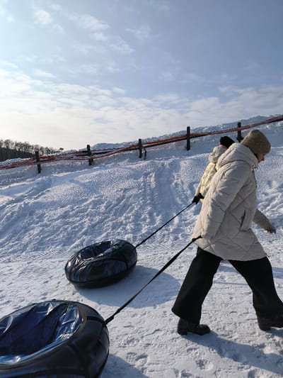 The image captures a winter scene on a snow-covered slope in Prybuzke, Ukraine, under a partially cloudy blue sky. A person, dressed in a light-colored puffer coat, dark trousers, and a light-colored beanie, walks from right to left across the frame, pulling two dark, patterned inflatable snow tubes uphill by black straps. A second person, partially visible behind the first, wearing a light-colored puffer jacket and a dark hat, is also ascending the slope. The slope exhibits visible tracks in the textured snow. In the background, a fence composed of dark posts and red-orange horizontal banding runs along the crest of the hill. A line of dark trees is visible on the distant horizon.