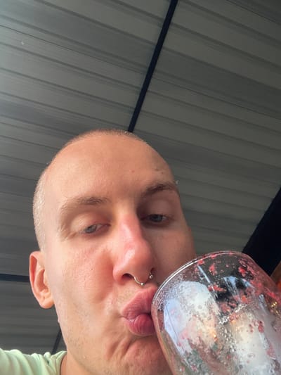 A close-up, low-angle shot depicts a person drinking from a glass. The person appears to be bald with fair skin, light-colored eyes, and a nose ring. The glass is tilted toward the viewer, revealing remnants of a red-colored beverage. The background consists of corrugated metal panels. There is no definitive information to suggest the location is Tambon Nam Phrae, Thailand.