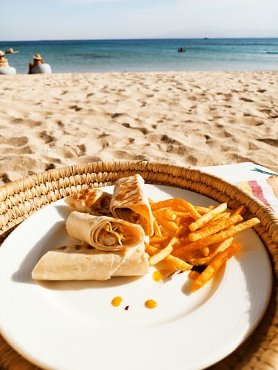The image depicts a white plate of food consisting of shawarma wraps and french fries, resting on a woven tray placed on a sandy beach. The scene is located in Ras Abu Soma, Egypt, indicated by the beach environment, the blue water of the Red Sea visible in the background, and figures sitting on the sand. The plate also has small dollops of sauce, a yellow mustard like one, and a reddish brown like ketchup. The tray holding the plate sits partially on a colorful striped towel. Beyond the tray, the sandy beach stretches towards the sea, where several figures are partially submerged in the water. A few people are seated near the shoreline on white beach furniture. The sky is clear and blue.