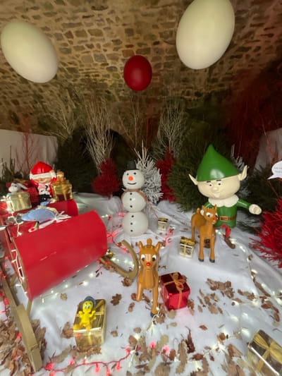 This image displays a static Christmas-themed exhibit situated within a stone-arched alcove in Bagnoles de l'Orne Normandie, France. The display's base consists of white fabric, which is covered with scattered dry, brown oak leaves and illuminated by multiple strands of warm-white LED string lights.
In the left mid-ground, a section of a red sleigh is visible, containing a Santa Claus figurine, several gold-wrapped gift boxes, and a light blue, cloud-shaped object. Centrally, a three-tiered white snowman figurine with a black top hat, orange carrot nose, and brown scarf stands upright. To its right, a green-clad elf figurine, wearing a pointed hat and tunic with a white collar, is positioned with its right arm slightly extended. Two brown reindeer figurines, each featuring a red nose, are located in the mid-ground: one stands near the elf, the other slightly forward and to the left.
Additional small gold and red wrapped gift boxes are distributed across the white base. A yellow Tweety Bird figurine is visible perched on a gold-wrapped box in the lower-left foreground. The background comprises a dense arrangement of artificial trees and branches in dark green, white, and bright red. The alcove's natural stone block ceiling features three suspended spherical decorations: two white and one red. A partially visible white speech bubble with indistinct text is present on the far right.