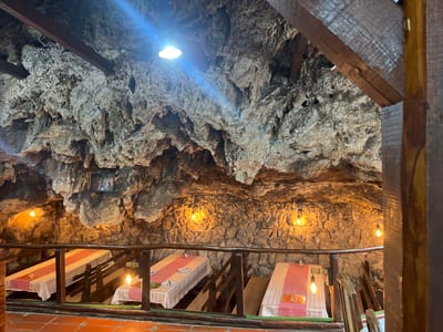 The scene depicts an unoccupied, multi-level indoor dining area with a natural cave or heavily cave-themed rock interior, located in Despotovac Municipality, Serbia, consistent with the Resavska Pećina Restaurant. The ceiling and upper walls exhibit irregular, coarse rock formations in mottled grey and brown tones, featuring prominent undulations and stalactite-like structures. A single bright, cool-white LED downlight is affixed to the ceiling, emitting a strong light ray. Below, approximately six warm, amber-toned Edison-style incandescent bulbs are suspended at varying heights from the rock walls, providing ambient illumination to the lower section. Dark wooden support beams are visible overhead, integrated with the rock ceiling. In the immediate foreground, a rustic wooden railing, comprising a rounded top rail and vertical posts, delineates a higher vantage point from a tiered seating arrangement below. This lower level contains multiple rectangular dining tables, each set with a white tablecloth and overlaid with either a red and white checkered pattern runner or a solid red runner. Some tables display plates and salt and pepper shakers. Longitudinal wooden benches accompany the tables. Wooden stairs facilitate access between the levels. A small, dark-colored plaque depicting figures in lighter tones is mounted on the left rock wall. No persons or overt activity are observed.