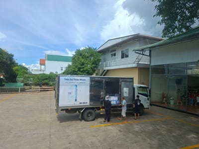 In Tân Phú 2, Vietnam, a white refrigerated truck is parked on a concrete lot, its rear doors open. Three men are engaged in unloading boxes from the truck. One man, wearing a hat and dark clothing, stands near the back of the truck, holding a box. Another man, in a light blue shirt and white shorts, stands to his right, also holding a box. A third man in a black t-shirt and shorts stands further to the right, near the truck's cabin. In the background, a building with a grey facade and a green roof is visible, along with trees and a cloudy sky. The truck has "TH True MILK" branding on its side. The area appears to be an industrial or commercial zone.