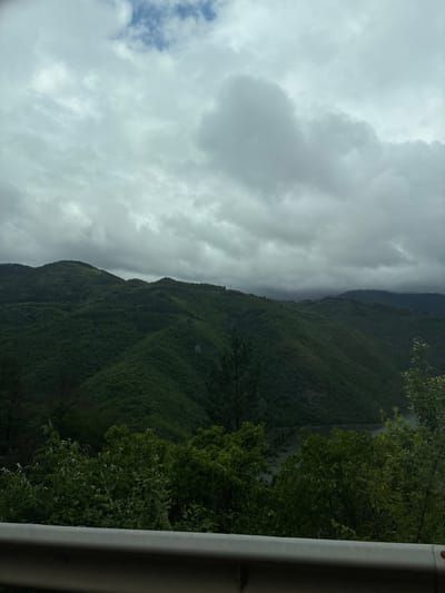 The image depicts a mountainous landscape under a cloudy sky, likely taken from a vehicle. Lush green vegetation covers the slopes of the mountains. A body of water, possibly a river or lake, is visible to the right of the mountains. A guardrail is visible in the foreground on the right side of the image. Based on the location provided (Liaskovo, Bulgaria), it is plausible the image depicts scenery near the Rhodope Mountains.