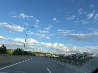 The image captures a highway scene under a bright blue sky with scattered white clouds. On the left side of the road, a concrete barrier runs parallel to the highway, with a line of trees behind it. Further in the background, rolling hills with scattered trees and what appear to be solar panels are visible. Several wind turbines are also present in the middle ground and background.
On the right side of the highway, a modern service area or rest stop complex is visible. This complex features a distinctive architectural style with angled, multi-level structures. Several businesses are advertised with illuminated signs, including "Wendy's" and "Subway." Cars are parked in front of the buildings. The highway itself has clear lane markings and a metal guardrail on the right side. The scene appears to be in a rural or semi-rural setting, likely near a major transportation route. The image does not depict the city of Tedotsminda, Georgia.