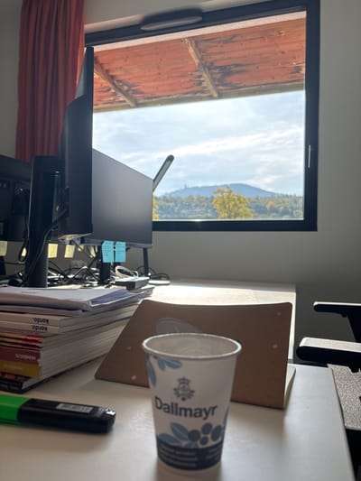 The image depicts an interior office workspace in Lièpvre, France, with sunlight streaming through a large window overlooking a natural landscape. In the foreground, a white paper cup branded "Dallmayr" in blue and white is centered on a white desk. To its left, a green and black highlighter rests horizontally. Further left, a stack of documents and books, some marked "kordo" and "numero 73," is present. A brown folder lies behind the cup.

The midground features a black computer monitor positioned on the desk, with cables and several blue sticky notes affixed behind it. Another monitor is partially visible to its left. A black articulating desk lamp is situated behind the primary monitor. A black office chair armrest is partially visible on the right side of the desk.

The background is dominated by a black-framed window showcasing an outdoor view. Beyond the window, rolling hills or mountains are covered with a mix of green and autumnal yellow-orange trees. A distinct structure, appearing as a tower or building, is visible on a distant hilltop. The sky is bright with scattered clouds. Above the window, an external wooden roof overhang is visible. To the far left, a section of an orange curtain hangs vertically. The interior walls are white.
