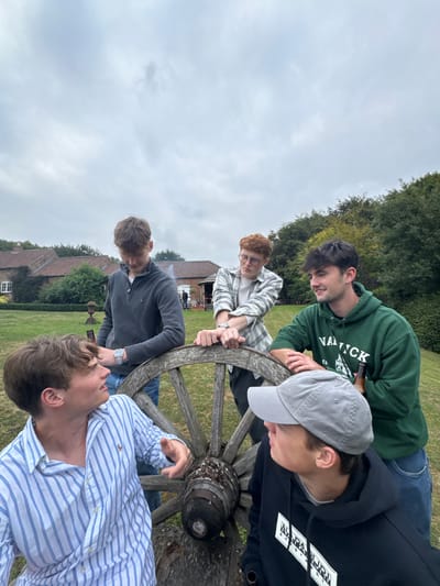 The image displays five young adult males gathered outdoors in a grassy area in Bishop Wilton, United Kingdom, under an overcast sky. A large, weathered wooden wheel, consistent with an antique wagon or cannon wheel, occupies the center.
From the left foreground, a male in a blue and white striped shirt faces right, his mouth slightly open, gesturing with his right hand toward a male in the immediate foreground right. This individual wears a grey baseball cap and a black hoodie with a white "NAPAPIJRI" logo, and is looking towards the first male.
Behind them, a male with light brown hair, wearing a dark grey quarter-zip top and blue jeans, leans his right arm on the wooden wheel, looking downwards. A silver watch is visible on his left wrist. To his right, a male with reddish hair, wearing glasses and a light-colored plaid shirt, also leans on the wheel, observing the male in the grey top. A silver watch is visible on his left wrist.
To the far right, a male with dark hair wears a green hoodie with a white "NAU TICK EST. 1989" logo. He leans on the wheel with his left arm and holds a brown bottle in his right hand, looking towards the right side of the frame.
The background features a well-maintained lawn, with traditional brick buildings featuring red tiled roofs visible further back. Dense green trees and foliage are present. A stone urn or planter is situated on the grass in the mid-ground, and a small wooden structure is visible to the right, with a faint plume of smoke or steam rising in the distance.