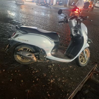 A white scooter is parked on a wet, gravelly road at night in Rhieng Blang, Indonesia. The scooter has gold rims and a carbon fiber exhaust cover. The rear tire is partially obscured by a metal bar in the foreground. In the background, other motorcycles and vehicles are visible, and people can be seen standing and riding. The scene appears to be illuminated by artificial lights, creating reflections on the wet road surface.