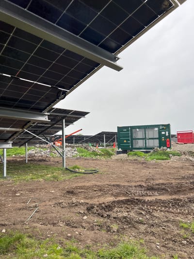 The image depicts an outdoor setting, likely a construction site for a solar energy installation in Grange Ville, Ireland. In the foreground, rows of solar panels are mounted on metal frames, their black surfaces reflecting the overcast sky. The ground is a mix of bare earth, tire tracks, and patches of green grass, suggesting recent construction or landscaping activity. In the middle ground, a green shipping container with safety symbols and text that includes "LV ROOM" stands near construction equipment, including an orange man lift or cherry picker. Further in the background, a red dumpster is visible. The overall impression is of an active solar farm construction project in a rural or semi-rural location.
