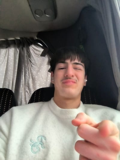 The image depicts a young male seated inside the passenger compartment of what appears to be a bus or large van. The specific location within Zlatina, Bulgaria, cannot be determined from the interior view.
**Person:** A male, likely in his late teens or early twenties, occupies the foreground, facing directly towards the camera. He has short dark hair with a fringe, dark eyebrows, a visible mustache, and fair skin with areas of lighter pigmentation around his mouth. He is wearing small silver hoop earrings in both ears and white wireless in-ear earbuds. His right eye (viewer's left) is closed in a wink, and his lips are slightly puckered.
**Attire:** He wears a light-colored, possibly off-white, long-sleeved knitted sweater. A light blue embroidered emblem, resembling stylized cursive letters, is visible on the left chest area of the sweater.
**Action:** The male extends his left hand towards the camera, with the index finger pointed directly at the viewer. His other fingers are curled inward.
**Environment:** The background indicates an enclosed vehicle interior. To the left, a light gray curtain with a subtle dotted pattern is drawn across a window. Behind the individual, a dark-colored seat with a textured or patterned fabric is visible. Above the subject's head, part of the vehicle's ceiling panel is visible, featuring two recessed circular elements, possibly lights or air vents. The lighting appears to be ambient, likely from external windows and possibly supplemented by interior vehicle lighting.