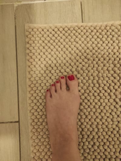 A person's right foot, with toenails painted red, is positioned on a textured, beige bath mat. The bath mat is placed on a tiled floor. The tiles are a light beige color with thin, darker grout lines. The scene appears to be indoors, likely in a bathroom. There is no identifiable location in Roeselare, Belgium, visible in the image.