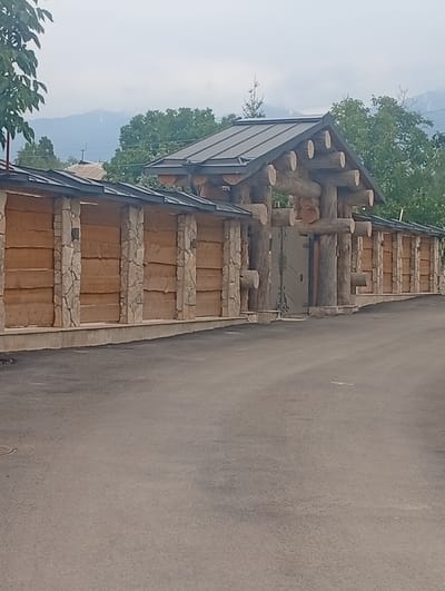 A rustic, heavy wooden gate with a metal arched insert is the focal point of this image, situated within a substantial fence constructed from large logs and stone pillars. The fence runs horizontally across the frame, with thick, stacked logs forming the main body of the wall, interspersed with vertical stone pillars that support the structure. The gate itself is framed by thick, rough-hewn logs, with smaller logs extending outwards horizontally, suggesting a sturdy, naturalistic design. A grey, metal gate with decorative scrollwork is visible behind the wooden frame. The fence and gate are set against a backdrop of lush green trees and a hazy sky with visible mountains in the distance, hinting at a mountainous or semi-rural location. The ground in front of the fence is paved with asphalt, showing signs of wear. The scene appears to be located in the city of Tash-Moinok, Kyrgyzstan.