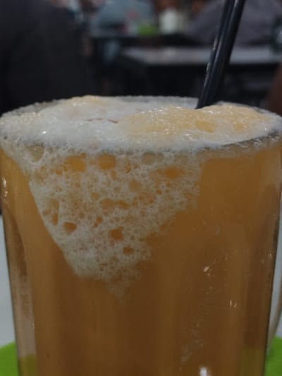 The image shows a close-up of a glass of frothy, orange-colored beverage with a black straw. The drink has a thick, foamy head with numerous small bubbles. The glass is sitting on a green surface. In the blurred background, there are other tables and people, suggesting a restaurant or cafe setting. Based on the prompt including the location of Santan, Indonesia, it is likely the drink is sold in that city.