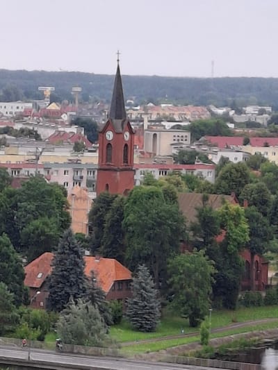The image depicts an elevated, wide-angle view of the city of Pila, Poland, likely captured from a distance. In the foreground, a bridge or elevated road runs horizontally across the bottom of the frame. Two figures on motorcycles are visible on this roadway. Behind the roadway, lush green trees dominate the middle ground, interspersed with a few buildings. A prominent brick church with a dark spire and clock faces stands out among the trees. Further beyond the church, the cityscape unfolds with numerous buildings of varying colors, including pastel pink, yellow, and grey, and some with red tiled roofs. In the background, a dense, dark green forest covers a hill, with a faint, tall structure, possibly a tower, visible on the horizon against an overcast sky. The overall impression is a panorama of an urban area surrounded by nature, under a cloudy sky.