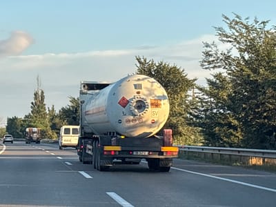 The image shows a tanker truck on a highway. The tanker is white with orange and red markings. It has a license plate that reads "BB-347-G". There are other vehicles on the highway, including a white van and a car. The highway is bordered by trees. The sky is blue with some clouds. The time appears to be late afternoon or early evening based on the lighting. The location is identified as the city of Mukhrani, Georgia.