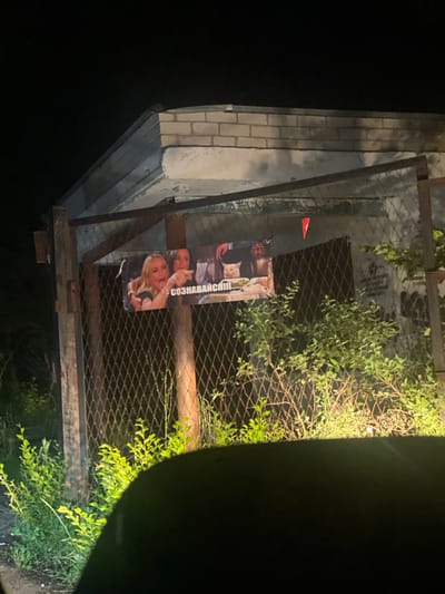 A poster depicting the "Woman yelling at a cat" meme is attached to a chain-link fence in front of a brick building. The poster features two women and a white cat sitting at a table. Text in Russian is visible on the poster, including "СОЗНАВАЙСЯ!!" which translates to "Confess!!". A red triangular flag is also attached to the fence above the poster. The scene appears to be outdoors at night, with some vegetation growing in front of the fence. The building behind the fence is made of light-colored bricks and has a weathered, possibly abandoned appearance. The foreground shows a dark area, possibly the interior of a vehicle, with green foliage illuminated by headlights. The image was taken in Ekaterinovka, Russia.
