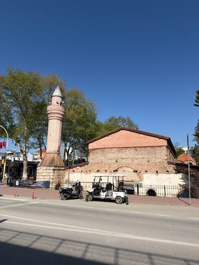 The image captures a sunny street scene in Iznik, Turkey, centered around historic architecture. On the left, a tall, cylindrical minaret constructed from reddish-brown bricks rises towards a clear blue sky, topped with a distinct conical roof. Large, leafy green trees partially frame the minaret and extend across the left side of the image.

Adjacent to the minaret, on the right, stands an old building with weathered brick walls and a red tiled roof, likely a mosque or another historical structure. Portions of its facade display exposed brickwork, while others are covered in lighter-colored plaster. Decorative metal railings are visible along the building, enclosing small arched windows.

In the foreground and stretching across the mid-ground is a paved street, marked with double white lines and a single yellow line. Several vehicles are parked along the right side of the road, including a white, open-sided vehicle resembling a golf cart and a black scooter or motorcycle. The far side of the street features more distant buildings, some appearing to be shops, identifiable by visible signs and Turkish flags. One prominent sign clearly displays "TÜRKCELL", a Turkish telecommunications company, and another reads "BURGER". A dark banner with Turkish text is also visible near street level, along with orange traffic cones.

The bright, cloudless blue sky and the strong shadows cast by the buildings and trees indicate clear, sunny weather during daylight hours, likely mid-morning or afternoon. The scene blends historical significance with elements of contemporary daily life in an urban setting.