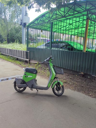 A green and grey electric scooter, labeled "B.bike", is parked on an asphalt path next to a wooden fence. Behind the fence is a green metal canopy with a transparent roof, under which a dark green car is parked. The car's roof and hood are visible. The area behind the canopy appears to be a residential building with windows. Trees and foliage are present in the background, and the sky is overcast. The scooter has a kickstand extended. A puddle of water is visible on the asphalt near the scooter. The scooter has a license plate or identification number on its front fender, reading "23-1470". The scene is likely in Putilkovo, Russia, based on the context provided.