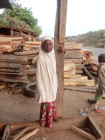 A young girl in a cream-colored hijab and a red and turquoise patterned skirt stands next to a wooden post. Her hand is holding the post. Behind her are stacks of lumber, a tire, and a wooden structure. To the right, a young boy in green pants and a patterned shirt is seen from the back. A road and a grassy hillside are in the background. The ground is dirt.