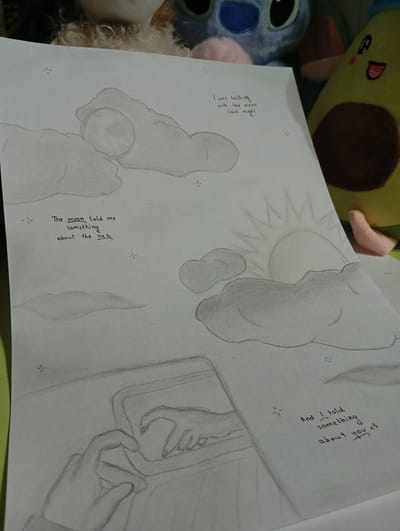 The image is a drawing on paper, likely a sketchbook page, depicting a celestial scene with a touch of anthropomorphism. In the upper left quadrant, a sketch of the moon is partially obscured by clouds. Below this, text reads, "The moon told me something about the sun". To the right, the sun is depicted rising or setting behind a cluster of clouds, radiating light. In the lower portion of the drawing, two hands are shown interacting with a device, possibly a tablet or phone, with one hand forming a heart shape. To the right of this, another line of text reads, "And I told something about you <3". The drawing style is pencil sketch with shading. The background of the photograph shows plush toys, including a blue Stitch character and a green avocado. The image is framed in a way that suggests it's placed on a flat surface, possibly a desk or table, with a bright light source casting shadows and highlights. There is no indication or visual information that would allow for the identification of the location as Kokand City, Uzbekistan.
