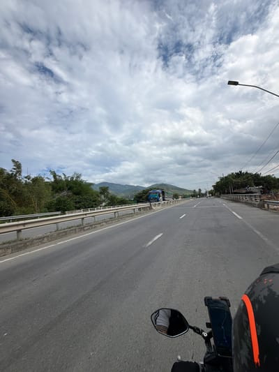 The image captures a first-person perspective from a motorcycle traveling on a road, as if viewed from the rider's right side. In the immediate foreground on the right, parts of the motorcycle are visible, including a black helmet with an orange stripe, a handlebar, and a smartphone mounted to the handlebar displaying a map application. A rearview mirror reflects a portion of the rider's torso and arm, clad in a white t-shirt.
The asphalt road, marked with white dividing lines, extends into the distance. A metal guardrail parallels the left edge of the road. Beyond the guardrail, a lush, green hillside covered with dense trees and vegetation descends. A blue truck is visible in the mid-distance on the road, partially obscured by the guardrail. Further in the background, a range of green mountains dominates the horizon.
The sky is heavily overcast with numerous white and gray clouds, though some patches of blue sky are visible. A street lamp, partially cut off, hangs from a pole in the upper left corner. The scene depicts a daytime journey through a scenic, mountainous, and forested landscape, indicative of a location like Cam Ranh Ward, Vietnam. The weather appears bright despite the cloud cover. No specific text is legible beyond the graphical interface of the navigation app.