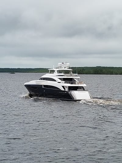 The image shows a large, modern yacht cruising on a body of water, likely a lake or wide river. The yacht has a dark blue and white hull with multiple decks and a radar or satellite dome on top. The name "DARIA" is visible on the stern. The water is choppy with the wake of the yacht clearly visible. The shoreline in the background is lined with trees and vegetation. The sky is overcast. No people are clearly visible in the image. The location is likely near a forested area with a body of water. The name "Gorushka, Russia" may be relevant, but cannot be confirmed with the information present in the image.