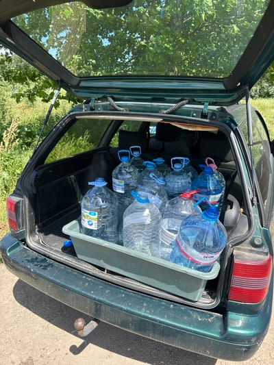 The open trunk of a dark green car is packed with a grey plastic bin filled with approximately 15 large, clear plastic water bottles. The location appears to be outdoors near foliage, possibly in Valchedram, Bulgaria. A tow hitch is visible on the car's rear bumper. The focus is on the filled water bottles in the car trunk. No people are visible.
