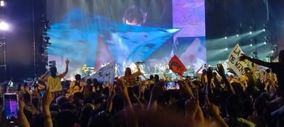 A vibrant night concert scene unfolds, filled with an enthusiastic crowd and a dynamic stage performance. The image captures the audience from a slightly elevated perspective, looking towards the brightly lit stage. A dense throng of people occupies the foreground and midground, with many raising their hands in excitement. Several attendees are on others' shoulders, while numerous smartphones are held aloft, recording the event.
On the left, a person in a yellow top stands on someone's shoulders, facing the stage with arms raised. Further into the crowd, another person waves a large, light blue flag or piece of fabric. A white banner is prominently displayed on the right, bearing partially visible black text that reads "LA DE MEND", likely part of "LA DE MENDOZA", suggesting a regional affiliation within Argentina. A red flag with an indecipherable white symbol is also visible.
The stage in the background is brightly illuminated with various colors, predominantly blues and purples. Large LED screens display dynamic visuals, including a performer with dark hair moving energetically. On the physical stage, several band members are visible, including what appears to be a drummer and a musician in a yellow top playing an instrument on the right. Tall sound and lighting towers flank the stage. The overall atmosphere is one of high energy and engagement, characteristic of a live music event in Tolosa, Argentina.