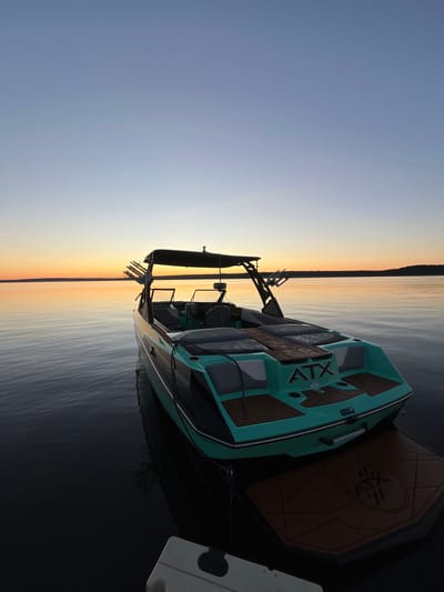 A teal and black wakeboard boat, branded with "ATX", is docked on a calm body of water at sunset in Gromovo, Russia. The boat is angled towards the right of the frame, with its rear facing the viewer. The water is dark and reflective, showing the subtle ripples of the gentle waves. A wakeboard tower with several boards attached is visible above the boat. The horizon line is low, with a gradient of soft orange, yellow, and blue hues from the setting sun painting the sky. The surrounding environment appears to be a tranquil natural setting with a distant tree line on the horizon. A portion of a white dock or platform is visible in the lower left corner of the image. There are no people visible in the scene.