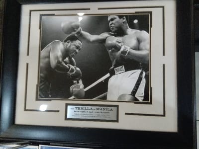 A framed black and white photo depicts Muhammad Ali throwing a punch towards Joe Frazier during their boxing match. The two boxers are inside the ring with ropes visible. Ali is wearing white shorts with "EVERLAST" on the waistband. A plaque beneath the photo reads "THE THRILLA IN MANILA, MUHAMMAD ALI - JOE FRAZIER". The frame is black with a white inner border.