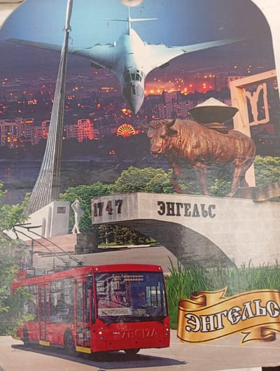 The image is a postcard depicting a collage of elements. In the background, a large bomber aircraft flies over a city at night, with illuminated buildings and a Ferris wheel visible. In the foreground, a red trolleybus is parked. To the right of the trolleybus, a golden ribbon displays text. Behind the trolleybus, there is a monument with a statue and the numbers "1747" and text that appears to say "ЭНГЕЛЬС". A sculpture of a bull is prominently placed on top of a structure. There are no people clearly visible in the image, other than indistinct figures inside the trolleybus. The image does not depict Khalkabad, Uzbekistan.