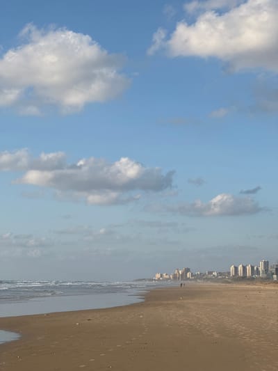 The image depicts a wide-angle view of the beach at Tirat HaCarmel, Israel, under a partially cloudy, light blue sky. The upper half of the frame is dominated by the sky, featuring scattered white and light gray cumulus clouds. The horizon line runs across the mid-section, separating the sky from the coastal landscape.
The left side of the lower half shows the Mediterranean Sea with moderate waves breaking onto the sandy shore, forming a white foamy edge. The water exhibits varying shades of grey-blue. The foreground and mid-ground consist of a wide expanse of light brown sand, showing numerous footprints and faint patterns.
In the mid-ground, two individuals are visible walking on the wet sand towards the distant cityscape, appearing as dark silhouettes against the brighter sand. A small, dark object rests on the sand to the left of their path.
Along the distant coastline, a series of multi-story buildings, likely residential, form a low-rise urban skyline. These structures appear light-colored and blend into the slight atmospheric haze, indicating a coastal city extending along the horizon. No immediate vegetation is prominent on the beach, but some distant, low-lying features are visible near the urban development. The scene is illuminated by natural daylight.