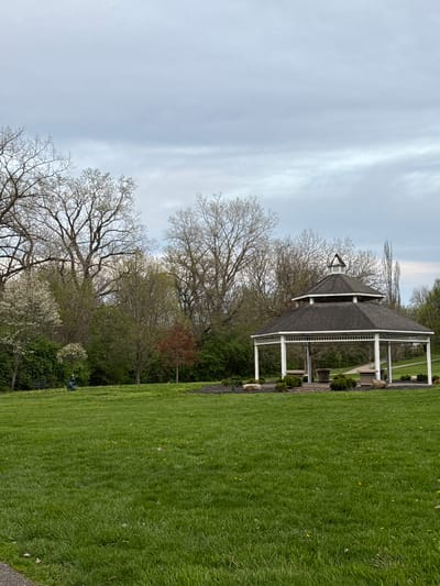 The image shows a gazebo in a grassy area. The gazebo has a dark grey roof, white supports, and is surrounded by trees. The sky is overcast. The location is likely a park in Grove City, Ohio.
