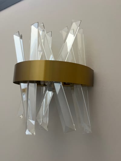 The image shows a close-up, eye-level view of a decorative wall sconce. The sconce features a band of brushed brass encircling a cluster of clear, angular acrylic or glass prisms. The prisms are arranged in a radiating pattern, with some extending upwards and others downwards, creating a geometric, starburst-like effect. The background is a plain, light-colored wall, likely painted. There are no people or other objects visible in the image. The lighting is soft and diffused, highlighting the reflective qualities of the prisms and the metallic finish of the brass band. The image does not provide any information about the location in Moni Community, Cyprus.