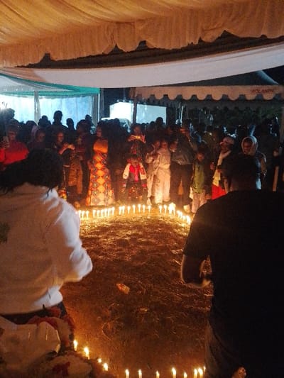The image captures an evening gathering in Marenyo-Shianda ward, Kenya, under a large fabric canopy. A prominent circular arrangement of numerous lit white pillar candles occupies the center of the bare earth ground. Within this candle ring, a young child is present, attired in a distinctive red, white, and black patterned outfit. Approximately 30-50 individuals, a mix of adults and children, surround the candle circle, observing the scene. Many spectators are actively holding smartphones, recording the event; one adult in the mid-ground wears a brightly patterned orange dress. In the immediate foreground, with their backs to the viewer, are two individuals: one wearing a white hooded top on the left, and another in a black t-shirt on the right. In the background, a white tent with "AOLANDIA" branding is partially visible, alongside other indistinct structures and ambient lighting.