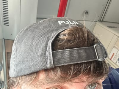 The media file is a close-up, eye-level shot of a person wearing a gray baseball cap that says "POLO" in white letters. The cap is positioned on the person's head, partially obscuring their hair, which appears to be light brown and slightly disheveled. The background suggests an indoor setting, possibly a mode of transportation, with a white, slatted panel on the left and a red bar running vertically in the upper center. The overall lighting is bright, indicating it's daytime. The visible portion of the person's face shows an eye with a green iris, and their skin tone is fair. There are no strong emotional cues conveyed by the image. The setting could be a train or bus interior, given the visible panels and the red bar, which might be a handrail. The focus is on the cap and the person's head, with the background blurred.