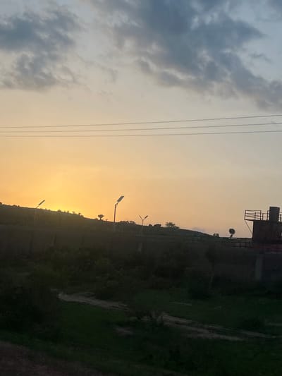 The image displays a landscape in Ribina East, Nigeria, captured during either sunrise or sunset, characterized by a prominent celestial gradient and silhouetted terrain. The sky transitions from a bright, warm orange-yellow near the horizon, indicating the sun's low position, to a muted blue-grey with scattered cumulus clouds in the upper regions. Three parallel utility lines are visible horizontally across the upper-middle portion of the sky.

A dark, elevated landmass or hill forms the horizon line, extending across the mid-section of the image. This terrain is sparsely dotted with the silhouettes of small trees and shrubs. Several artificial structures are present on this elevated ground: two distinct streetlights featuring rectangular solar panels are silhouetted, one on the left and another positioned more centrally to the right. A smaller, conventional streetlight silhouette is also visible further to the right. On the far right, a dark, geometric structure, possibly a water tower or similar industrial installation, stands with an elevated platform and railings.

The foreground occupies the bottom third of the image and is heavily shadowed, appearing as dense, dark vegetation consistent with overgrown fields or undeveloped land. Subtle variations in tone suggest uneven ground and potential dirt paths within the dark foliage. No individuals or discernible actions are present within the scene.