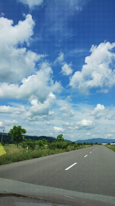 The image depicts an asphalt road stretching into the distance, bordered by lush green vegetation and rolling hills under a bright blue sky filled with fluffy white clouds. The road has a dashed white line marking its center. The landscape appears to be rural, with scattered trees and fields visible along the sides of the road. In the distance, mountains can be seen. The photograph seems to be taken from a vehicle traveling on the road. The location is Velyatyno, Ukraine.