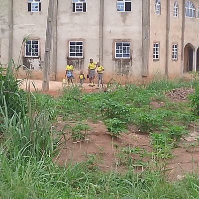 The image shows four children in school uniforms, standing in front of a building with a beige facade. They are all wearing yellow shirts with blue accents and are carrying backpacks. The building has several windows and a door, and there are some plants growing in the foreground, in front of the building. The scene takes place in Adanisievbe, Nigeria, located at the coordinates 5.955 latitude and 5.704 longitude. The children are all looking at the camera and appear to be in a good mood. The environment appears to be a rural setting with a lot of green plants and some dirt ground. The children are likely on their way to or from school.