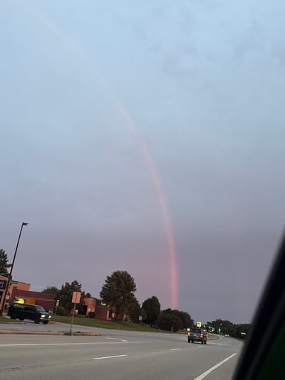 The image depicts a scene on a multi-lane road in Bloomingdale, United States of America, with a faint rainbow visible in the overcast sky. In the foreground, the edge of a vehicle is visible on the right side. The road has visible lane markings and is bordered by a median with trees and grass.
On the left side of the road, a black van is parked, with text on its side reading "PATRICK POOL & SPA". Further back, a commercial building with brick accents is visible, along with trees and a streetlamp. Several cars are driving on the road, including a black SUV and other vehicles further in the distance. Traffic lights are visible in the distance, indicating an intersection. The overall lighting suggests it is either dusk or dawn, with a soft, diffused light. The sky is a pale gray with subtle hues of pink and purple, likely due to atmospheric conditions. The rainbow appears as a soft, vertical arc of reddish-pink light.
