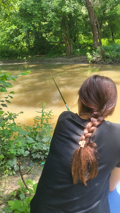 The image captures an outdoor scene in the city of Vishna, Bulgaria, featuring a person fishing by a calm, murky yellowish-brown river or large stream under bright daylight conditions. The individual is viewed from behind, wearing a plain black short-sleeved t-shirt. Their long, dark reddish-brown hair is styled into a single, textured braid down their back, secured near the end with a light brown claw clip. They are holding a slender, dark green fishing rod with an orange-tipped float, extended into the water where the fishing line is visible. The person is seated on a blue object, likely a chair or cushion, partially visible at the bottom right. The near bank, a muddy earth shoreline, is overgrown with various green plants, small bushes, and ground cover, with some small white flowers discernible. Scattered green and blue debris is also present on this bank. The opposite bank is densely forested with mature deciduous trees and lush undergrowth, their reflections visible on the water's surface. A large fallen tree branch lies partially submerged or on the far bank. Numerous small, white particulate specks, possibly pollen or insects, are visible in the air throughout the sunlit background.