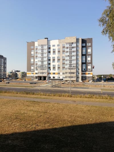 A multi-story apartment building under construction is pictured in Zhabinka, Belarus. The building has a modern design with a facade that combines grey, white, and black sections, as well as some beige and orange accents. It features numerous windows and balconies. A construction crane is visible on the right side of the building, with a person in a red gondola suspended from it. In the foreground, there is a dry grass field, a paved walkway, and a road. Other buildings, some also under construction, are visible in the background, along with some trees and a clear blue sky. The overall scene suggests an active construction site in a suburban or newly developed area.
