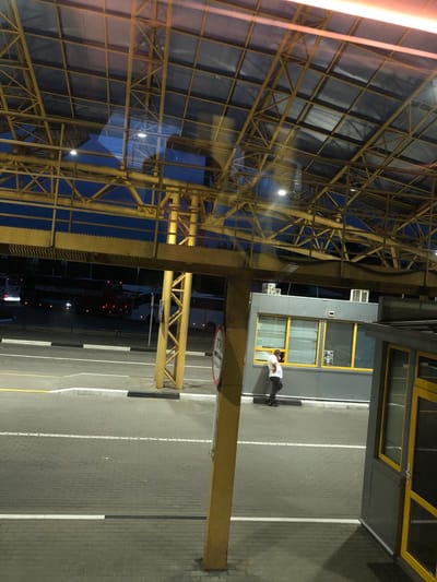 The image shows an outdoor scene at what appears to be a border crossing or transportation hub in Starovoitove, Ukraine. A man in a white t-shirt and dark pants is standing near a grey building with yellow trim. He is facing away from the camera, looking towards the building. Two air conditioning units are mounted on the exterior of the building. The ground is paved with grey bricks and marked with white and black lines, indicating lanes or pedestrian walkways. In the background, several buses are visible, some with their lights on. Above, a large yellow metal structure with a transparent roof is visible, with reflections of the sky and lights on the roof. A yellow pillar supports part of this structure in the foreground. A red and white circular sign with text is partially visible on the right side of the image. The lighting suggests it is dusk or night.